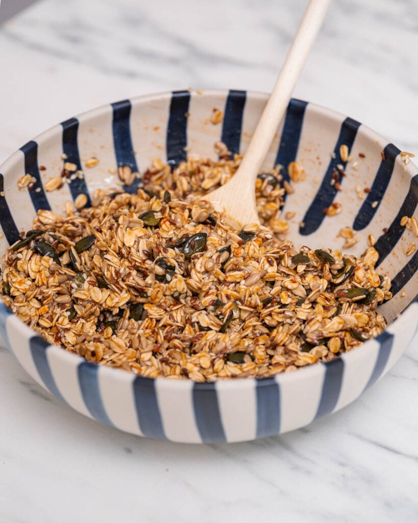 A bowl of granola with seeds and nuts, mixed with a wooden spoon, sits on a marble countertop.