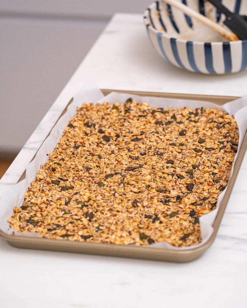 Ein Backblech voller Granola Salé Maison steht auf einem mit Backpapier ausgelegten Blech auf der Marmorarbeitsplatte. Im Hintergrund runden eine Keramikschüssel und Holzutensilien diese rustikale kulinarische Szene ab. 