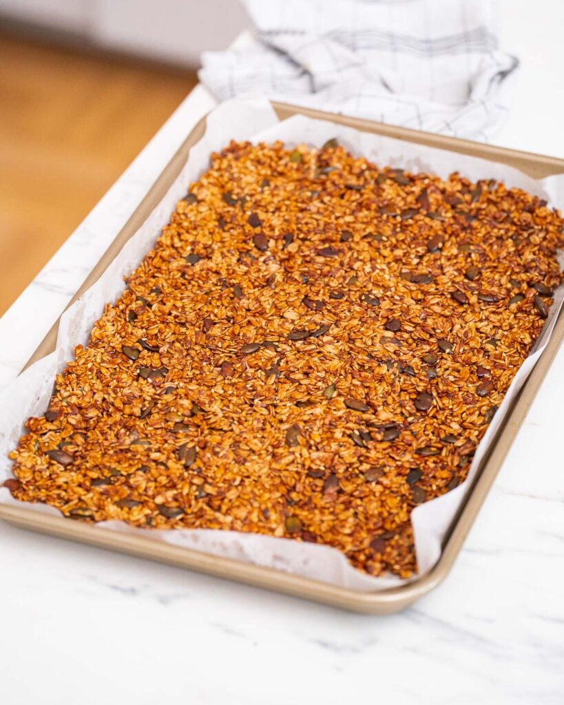 A baking sheet lined with parchment paper holds freshly baked savory granola, made from oats and seeds, placed on a white countertop.