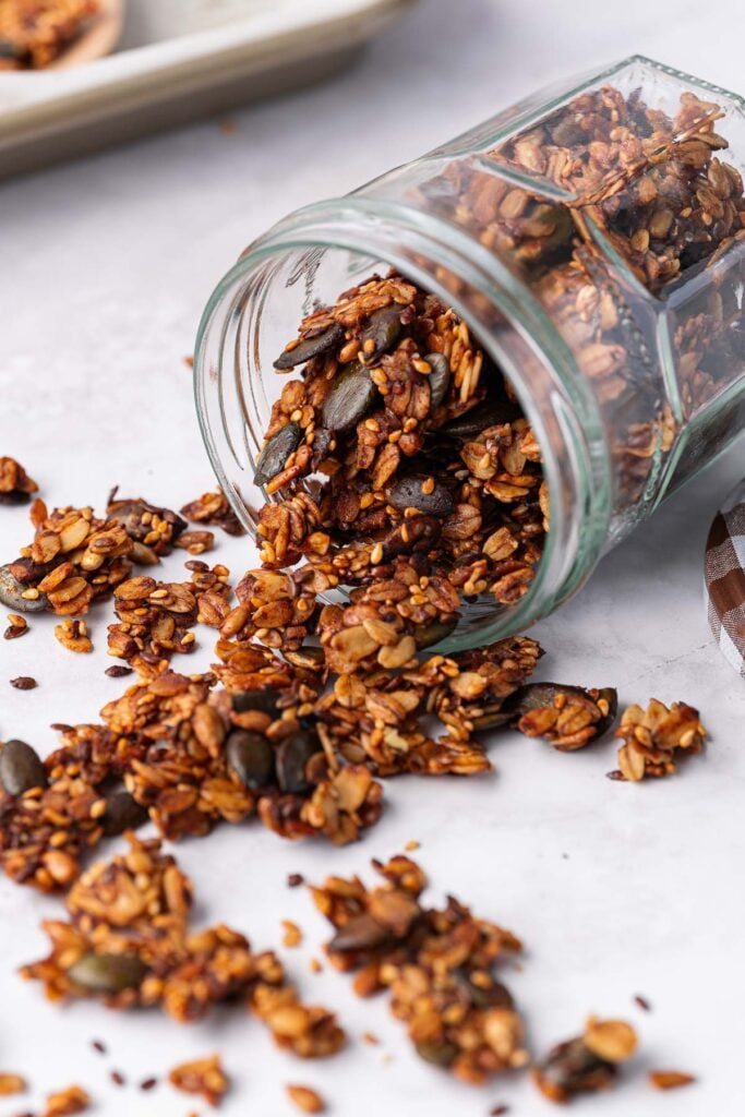 The jar tipped over, spilling a savory granola mix with seeds and nuts onto a white surface.