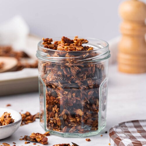 Ein Glas mit herzhaftem Granola steht auf einer Marmorplatte. Daneben liegt ein Löffel mit weiterem Granola, bereit zum Genießen. Im Hintergrund sorgen eine Pfeffermühle und ein Backblech für eine rustikale Note.