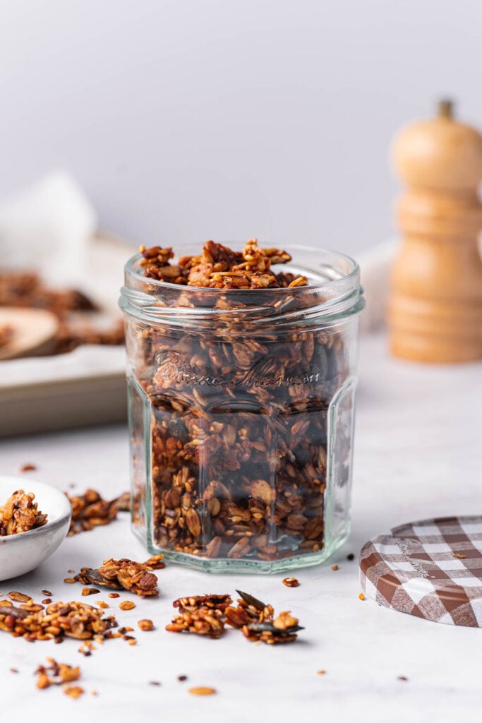 A jar filled with savory granola sits on a marble countertop. A lid is nearby and a blurred pepper mill appears in the background. 