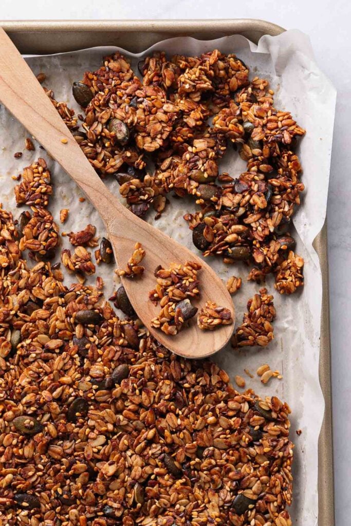 A baking tray with homemade savory granola made from a variety of seeds and cereals. A wooden spoon rests on the baking sheet, surrounded by scattered pieces of granola. 