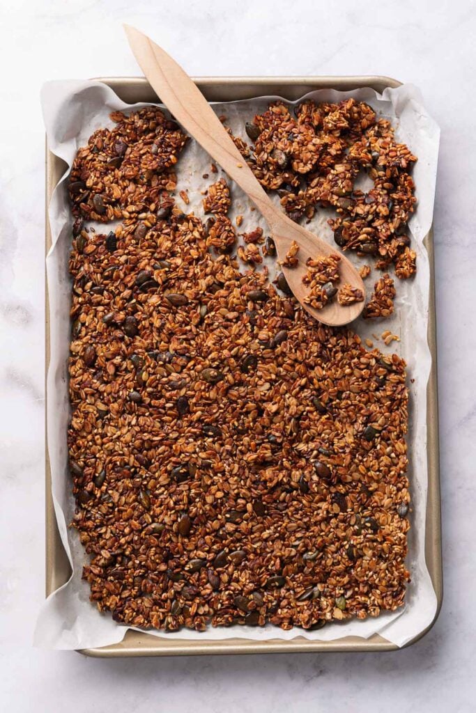 Gebackenes Müsli mit einem Holzlöffel auf einem mit Backpapier ausgelegten Backblech verteilen.