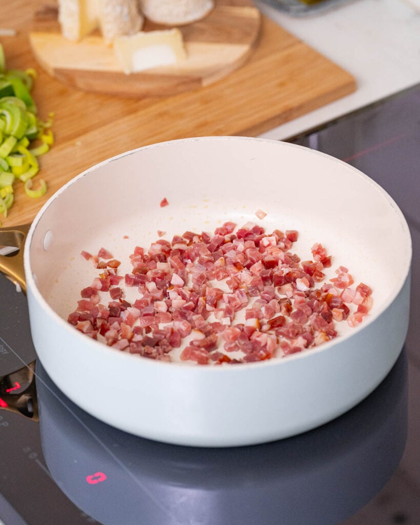 Lardons coupée en dés cuite dans une poêle blanche sur une cuisinière à induction avec des poireaux hachés sur une planche de bois en arrière-plan.