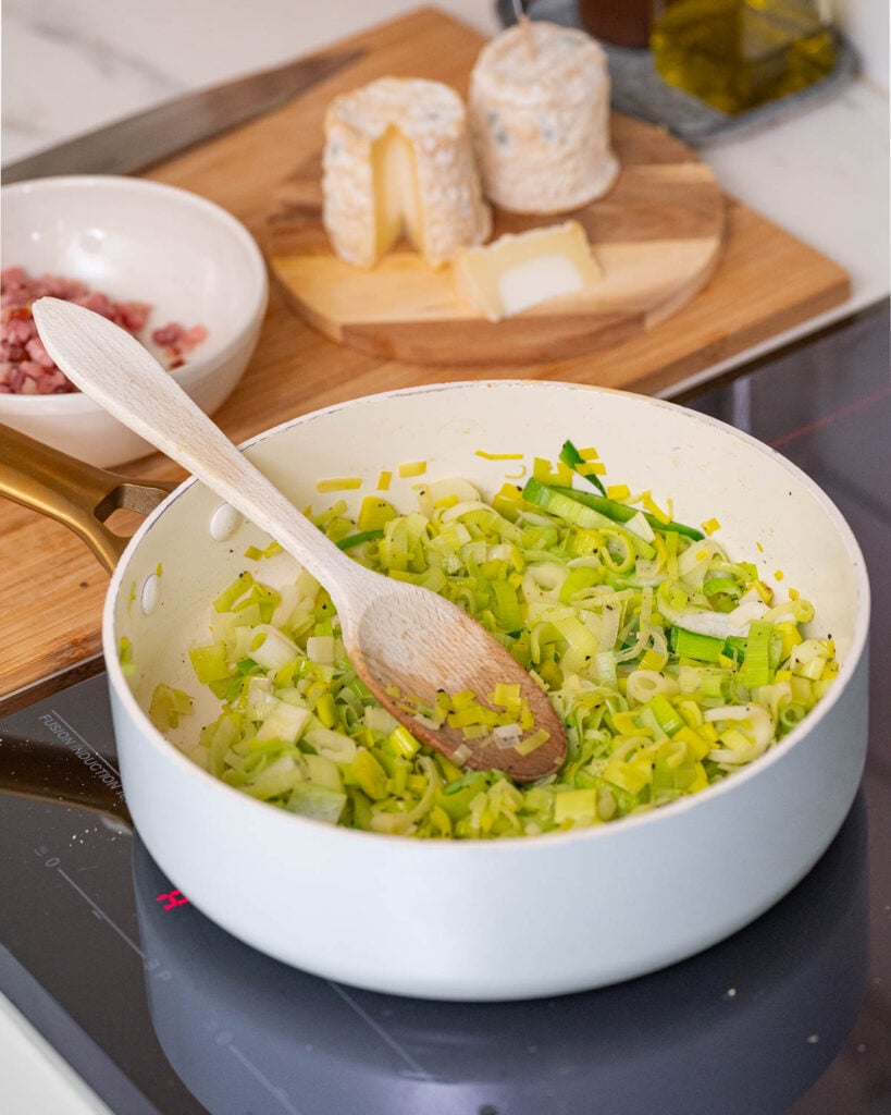 Une poêle blanche sur une cuisinière contient des poireaux sautés avec une cuillère en bois. En arrière-plan, il y a une planche à découper avec du fromage de chèvre et un bol de lardons.