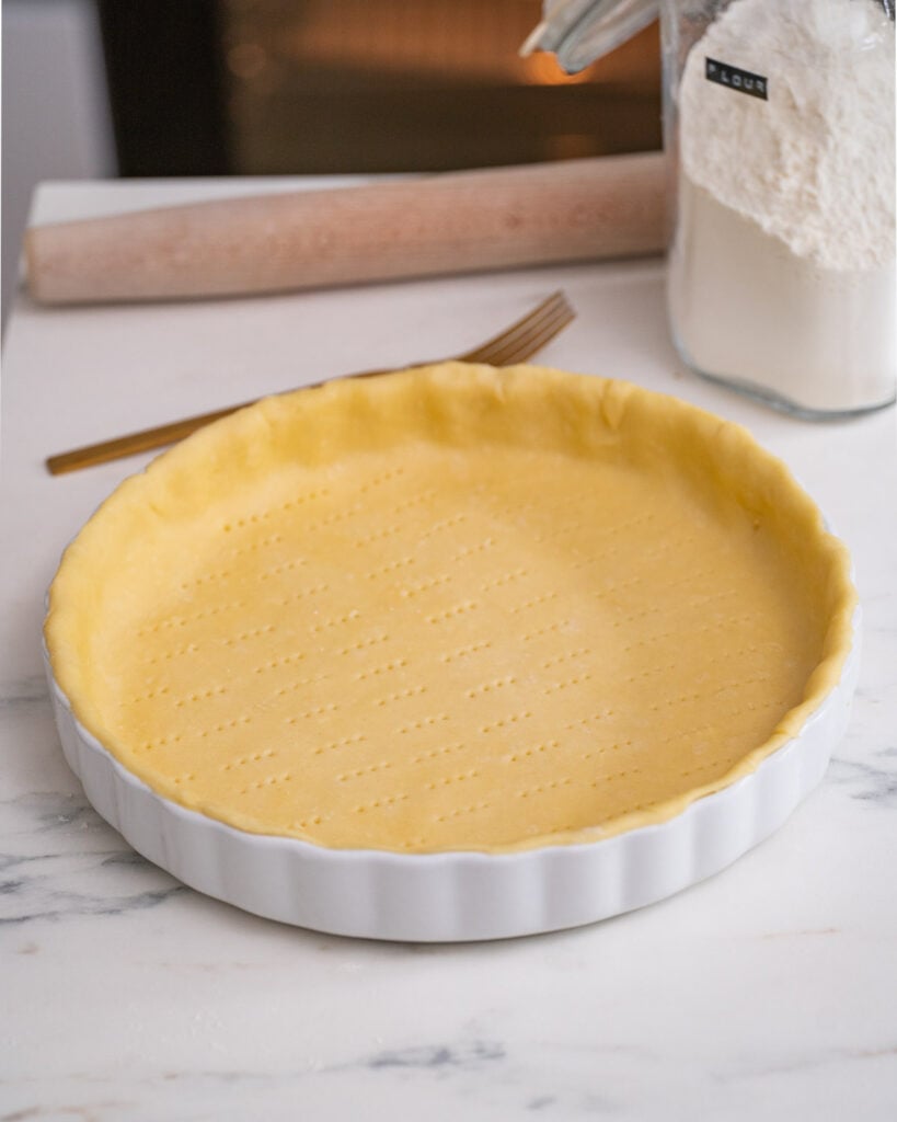 A shortcrust pastry pricked with a fork to prepare a leek-goat-lardons quiche on a marble work surface.