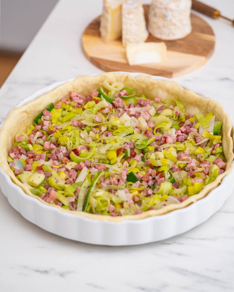 An unfired shortcrust pastry topped with chopped leeks, diced bacon and slices of goat's cheese in a white ceramic dish on a marble surface, reminiscent of a classic leek quiche.