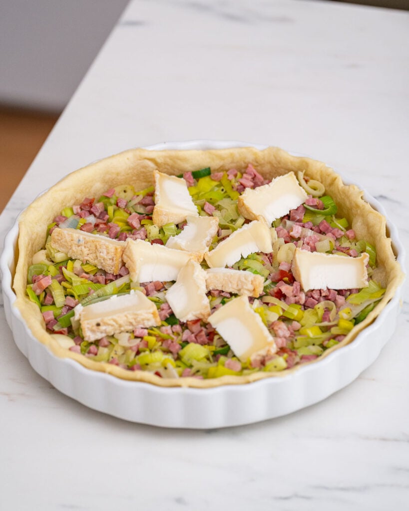 Une pâte brisée non cuite garnie de poireaux hachés, de lardons en dés et de tranches de chèvre dans un plat en céramique blanche sur une surface en marbre, rappelant une quiche aux poireaux classique.