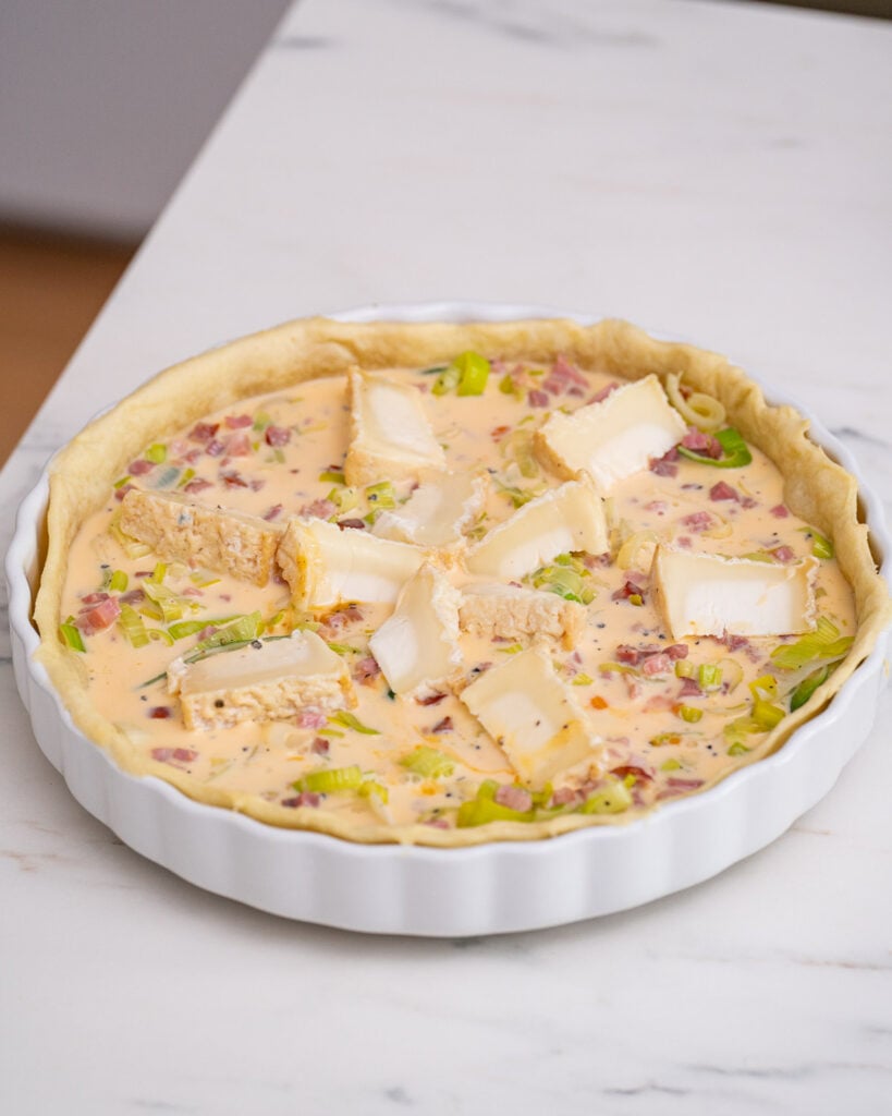 Raw leek and goat cheese quiche in a white dish, topped with a creamy mixture, diced vegetables, slices of cheese and bacon.