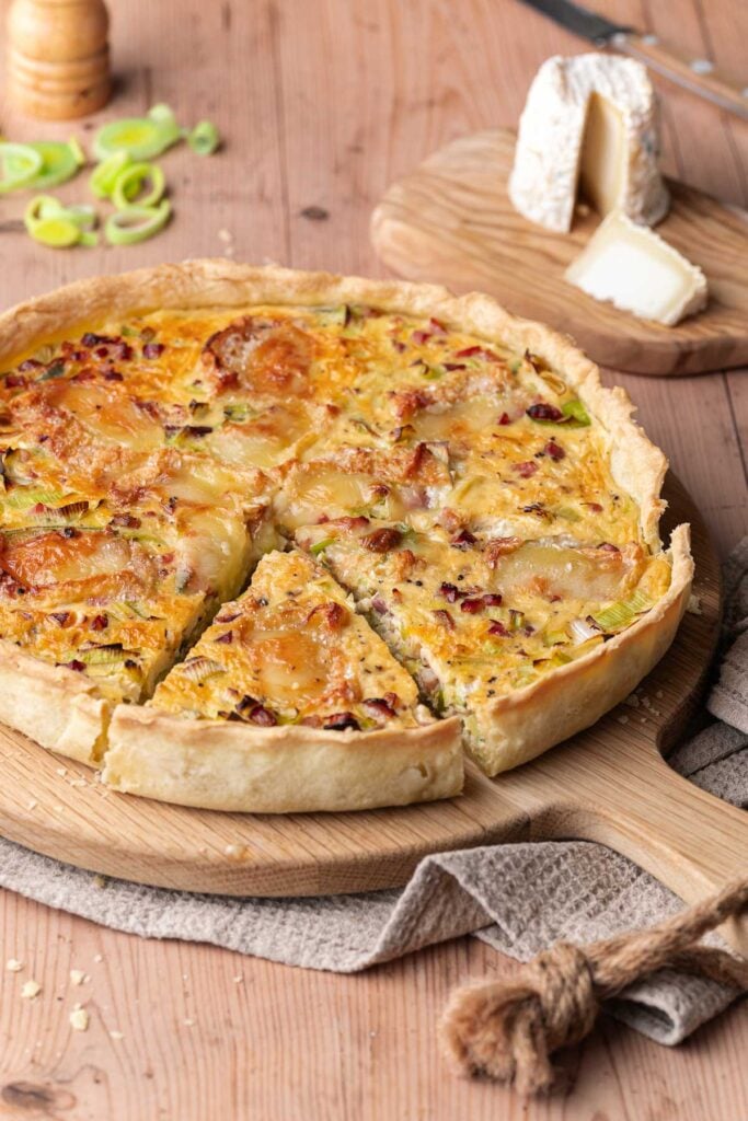 A sliced leek, goat's cheese and bacon quiche rests on a wooden board, with fresh leeks and creamy cheese in the background.