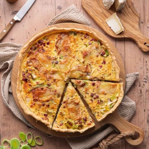 A leek-goat quiche with one slice removed rests on a wooden plank. Nearby, cheese, a knife, sliced leeks and a pepper mill adorn the rustic wooden table.