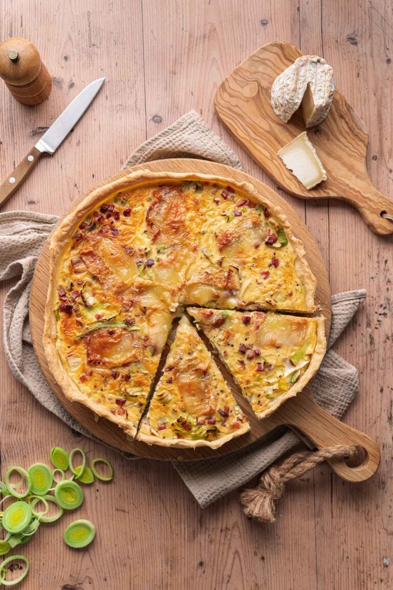 A leek-goat quiche with one slice removed rests on a wooden plank. Nearby, cheese, a knife, sliced leeks and a pepper mill adorn the rustic wooden table. 