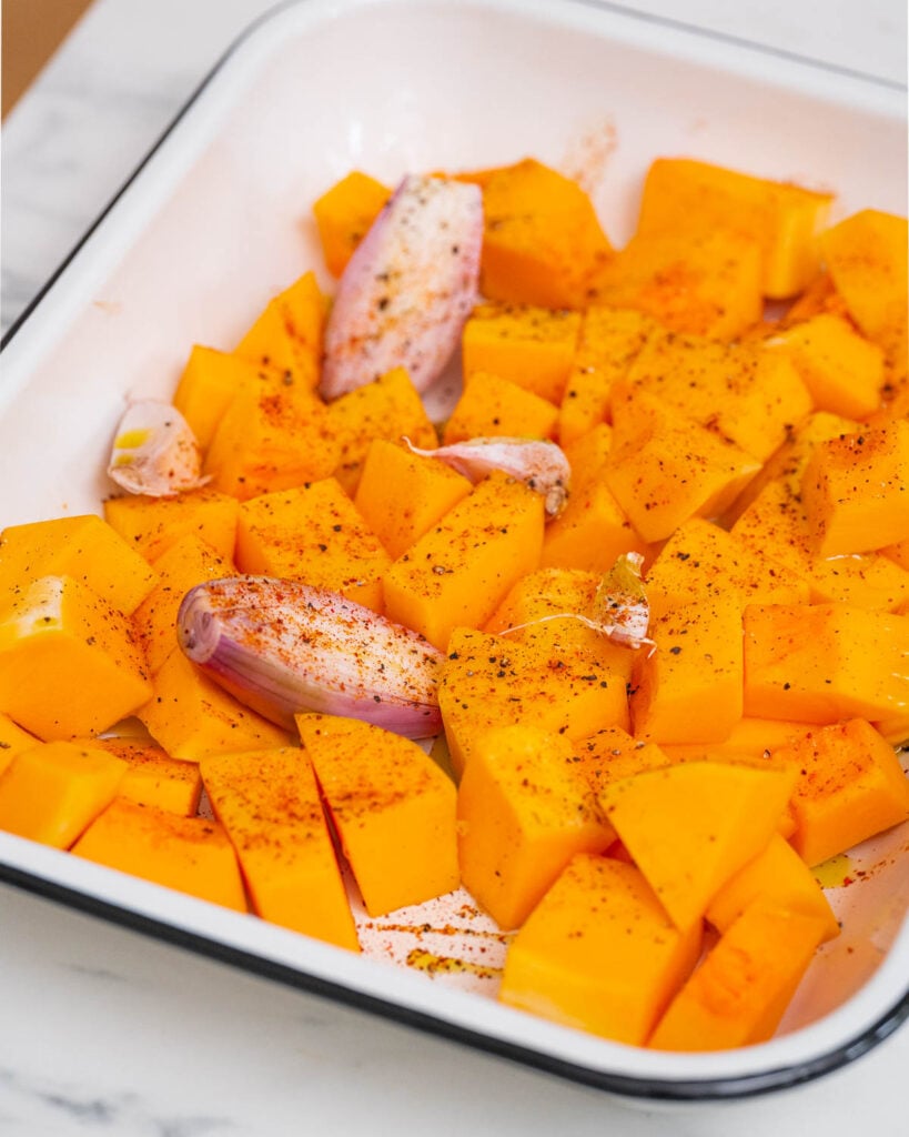 Chopped butternut squash and garlic cloves, seasoned with spices, create a delicious butternut velouté in a white baking dish.