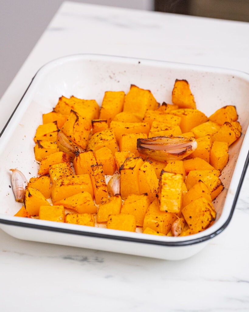 Roasted butternut squash cubes and garlic cloves are elegantly arranged in a white baking dish on a marble surface, reminiscent of creamy butternut velouté.