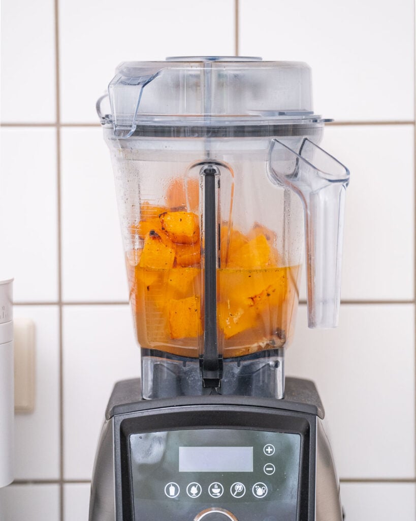 Ein Mixer mit gehackten Orangenfrüchten und Noten von Butternut-Suppe steht auf einer Küchentheke an einer gefliesten Wand.