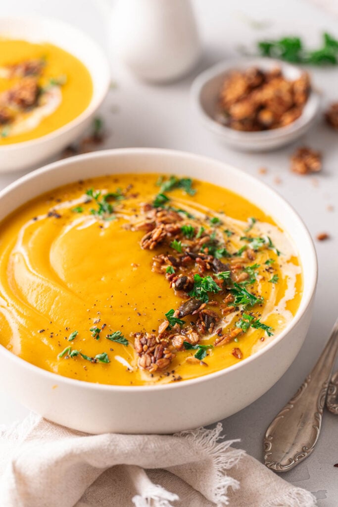 A bowl of creamy butternut velouté garnished with pecans and parsley, with a swirl of cream. A napkin and spoon are placed next to the bowl, while another bowl and pecans are cleverly blurred in the background. 