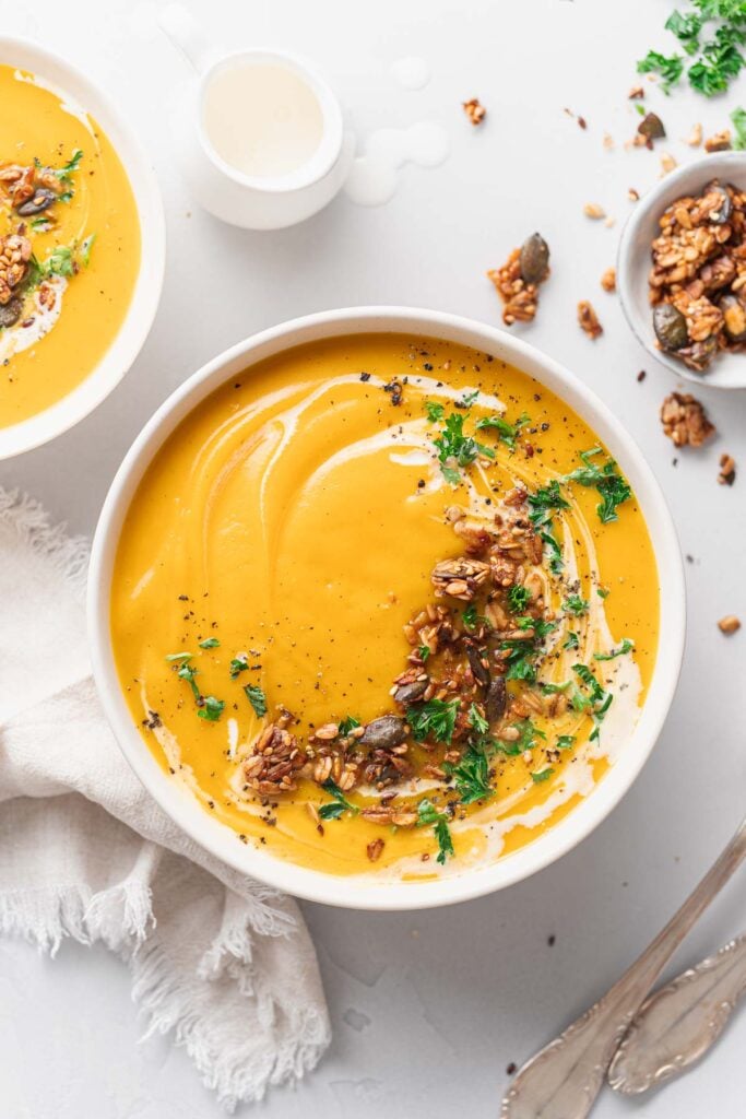 Eine Schüssel mit cremiger Butternut-Suppe, garniert mit Samen, Nüssen, Kräutern und schwarzem Pfeffer. Ein kleiner Topf mit Sahne und ein weißes Tuch stehen in der Nähe, neben einer weiteren Schüssel mit Nüssen und Samen. 