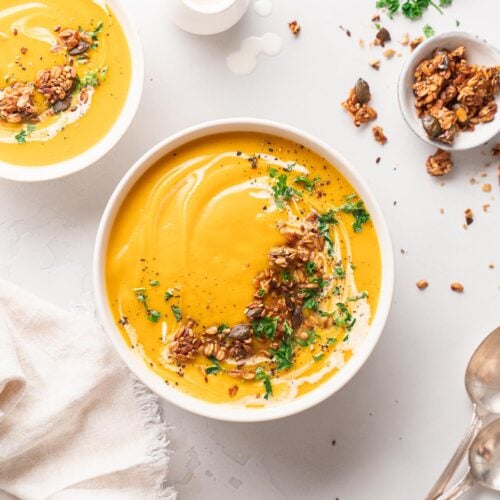 Drei Schalen mit cremiger Butternut-Suppe sind mit Kräutern und Samen garniert. Ein kleiner Krug und Silberlöffel werden in der Nähe auf einer hellen Oberfläche platziert.