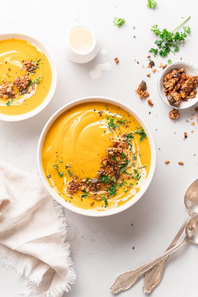 Two bowls of butternut velouté are garnished with cream, seeds and herbs. A small dish of granola, a cream pitcher and two silver spoons rest nearby on a white surface covered with a tea towel. 