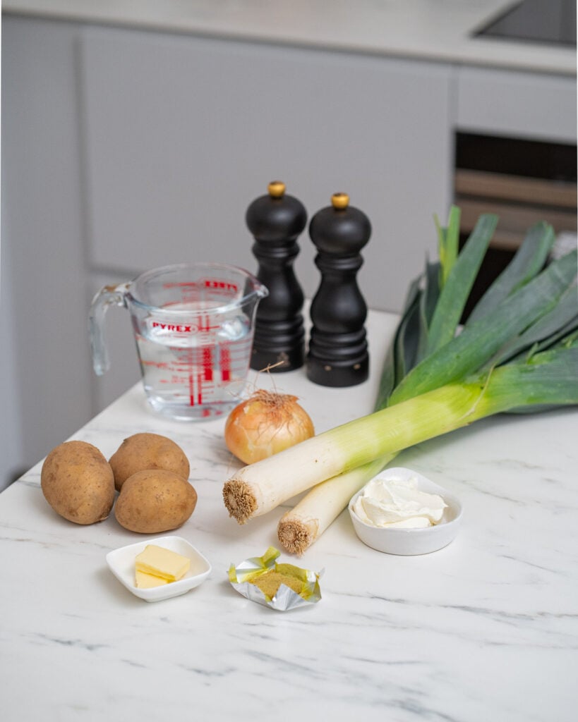 Ingredients for countertop leek and potato soup: leeks, potatoes, onion, butter, cream, bouillon cubes, water in measuring cup and salt and pepper mills.