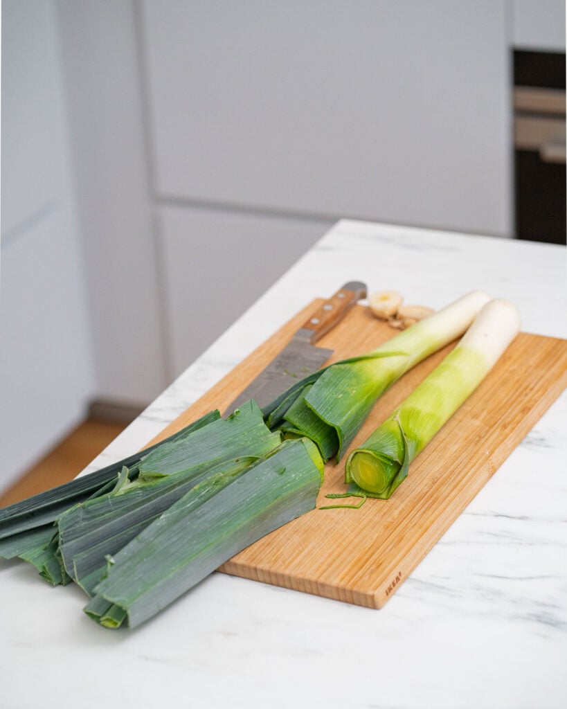 Chop leeks whole with a knife on a wooden cutting board on a kitchen counter.