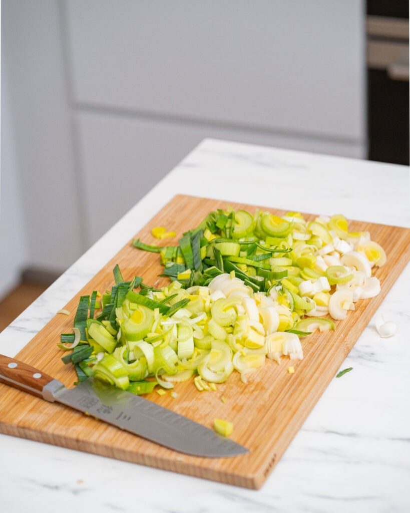 Poireaux hachés sur une planche à découper en bois à côté d'un couteau, posé sur un plan de travail de cuisine blanc.