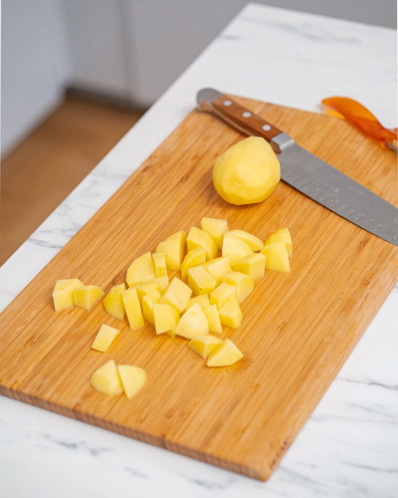 Chopped and whole potatoes peeled on a wooden cutting board with a knife and peeler.