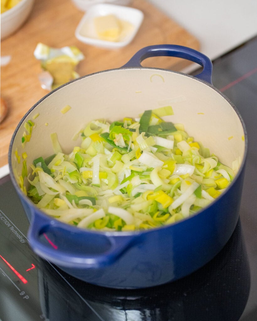 Poireaux hachés en train de cuire dans une casserole bleue sur une cuisinière, avec des ingrédients en arrière-plan.