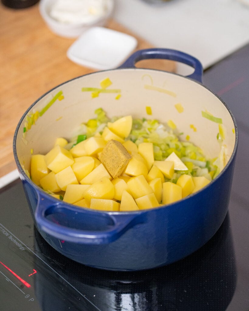 In einem blauen Topf auf dem Herd befinden sich gehackte Kartoffeln, Lauch und ein Brühwürfel.