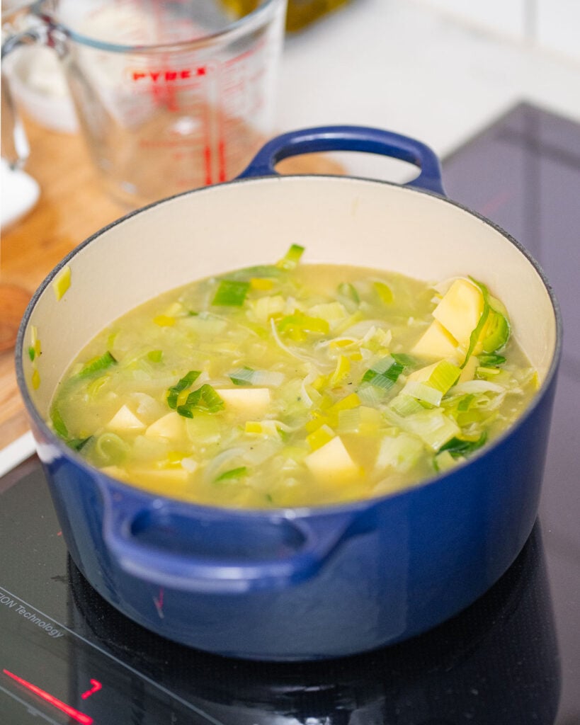 Une marmite bleue contenant de la soupe aux poireaux hachés et des morceaux de pommes de terre mijote sur une cuisinière à induction. Une tasse à mesurer est visible à l'arrière-plan.