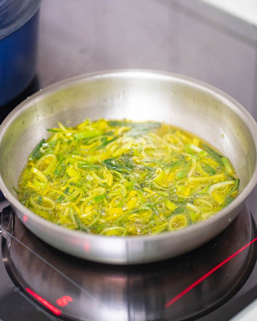 A skillet on a stovetop with sliced greens, probably leeks, sizzling in the oil, reminiscent of the beginnings of a delicious leek potato soup.