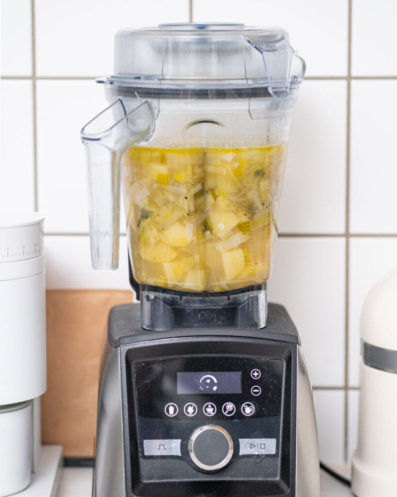 A blender filled with chopped vegetables and liquid is placed on a kitchen counter against a white tiled wall.