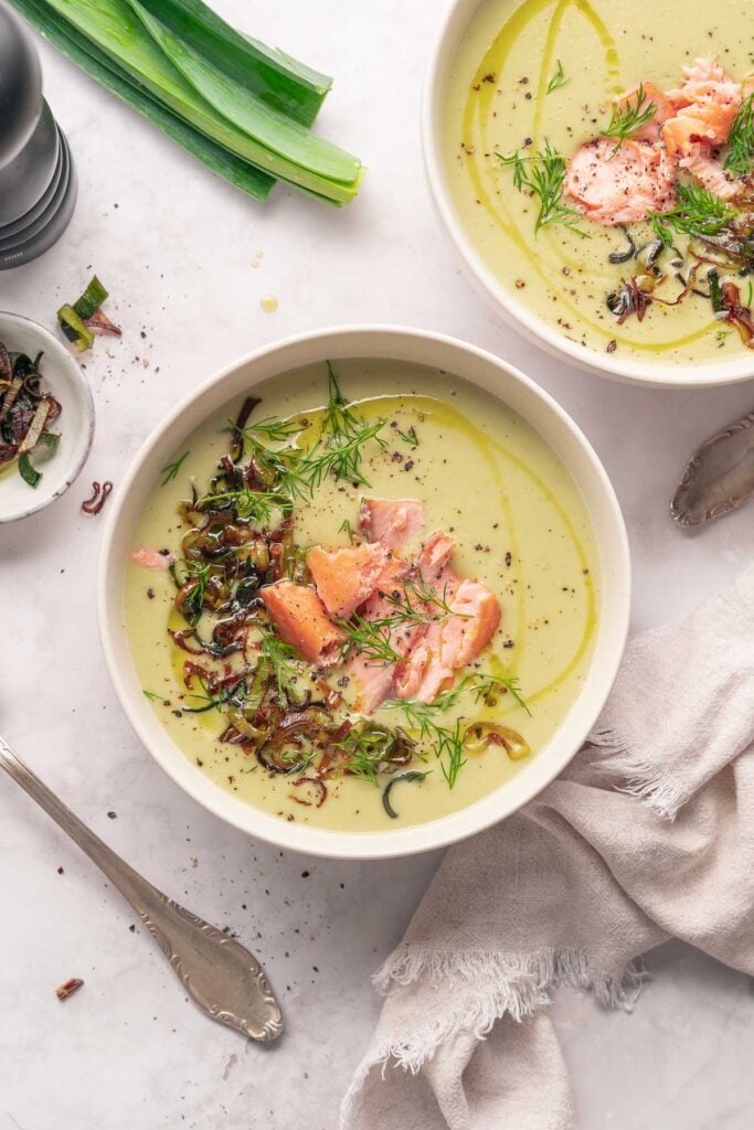 Un bol de soupe crémeuse aux poireaux et pommes terre garnie de saumon, d'aneth et de légumes sautés repose à côté d'une cuillère et d'une serviette sur une surface en marbre.