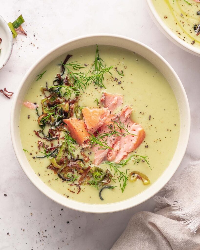 A bowl of creamy green soup topped with chunks of salmon, sautéed leeks and fresh dill on a light background.