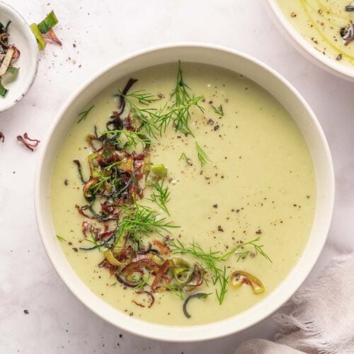 A bowl of creamy leek and potato soup garnished with chopped onions, fresh dill and cracked pepper, placed on a light surface with a towel beside it.