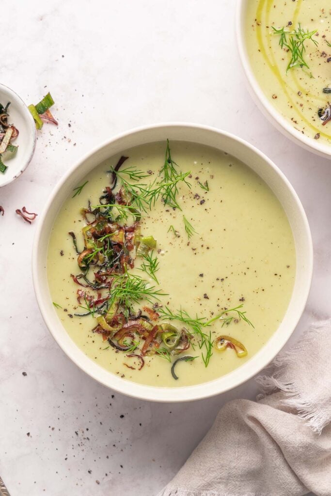 A bowl of creamy green soup garnished with chopped onions, fresh dill and cracked pepper, placed on a light surface with a towel beside it.