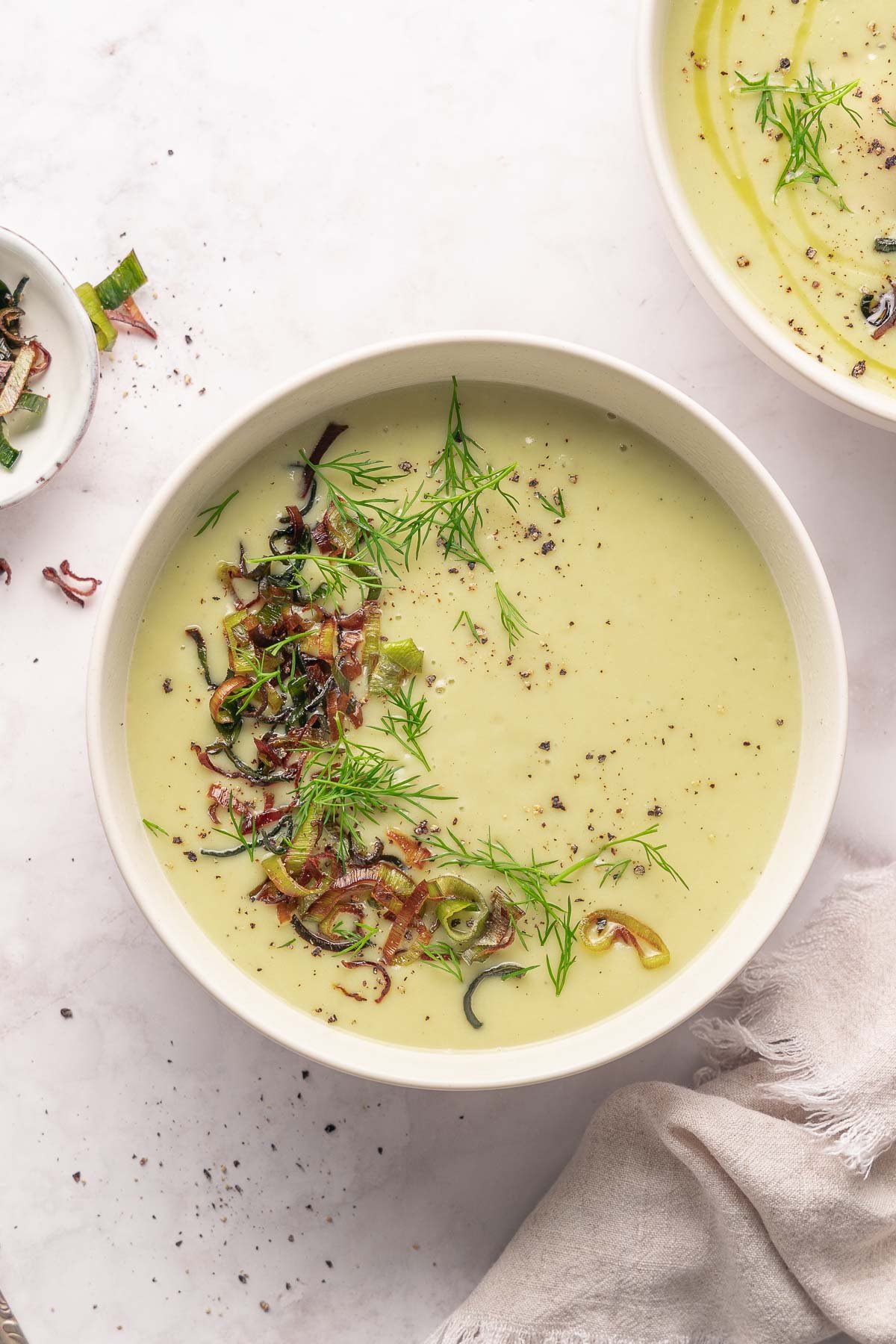 Eine Schüssel cremige Lauch- und Kartoffelsuppe mit gehackten Zwiebeln, frischem Dill und zerstoßenem Pfeffer, auf einer hellen Oberfläche mit einem Handtuch daneben platziert.