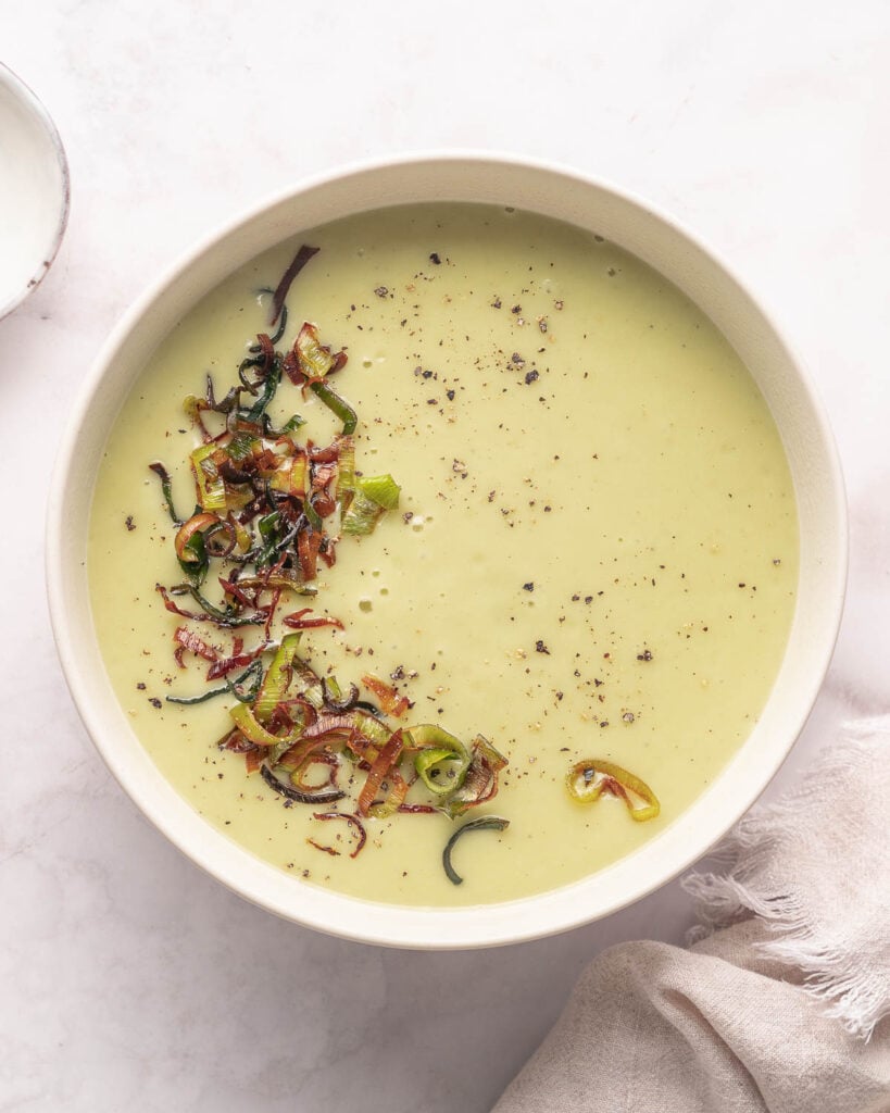 A bowl of creamy soup topped with crispy fried vegetables and black pepper, placed on a light-colored surface.