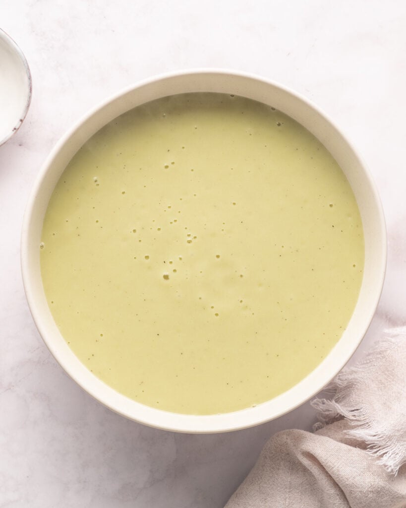 Un bol de soupe lisse, vert clair, sur une surface blanche avec un tissu beige partiellement visible.