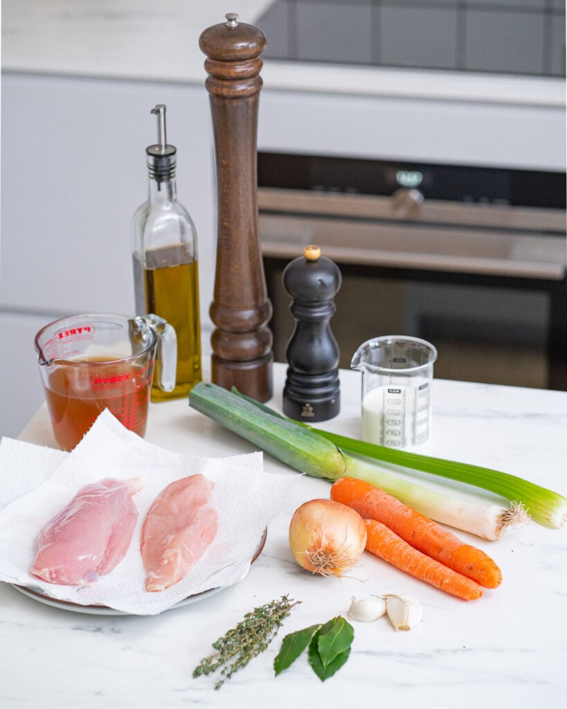 Ingredients for Creamy Vegetable Chicken Soup: raw chicken breasts, carrots, onion, leek, garlic, thyme, bay leaves, olive oil, broth, milk and salt and pepper mills on a kitchen counter.