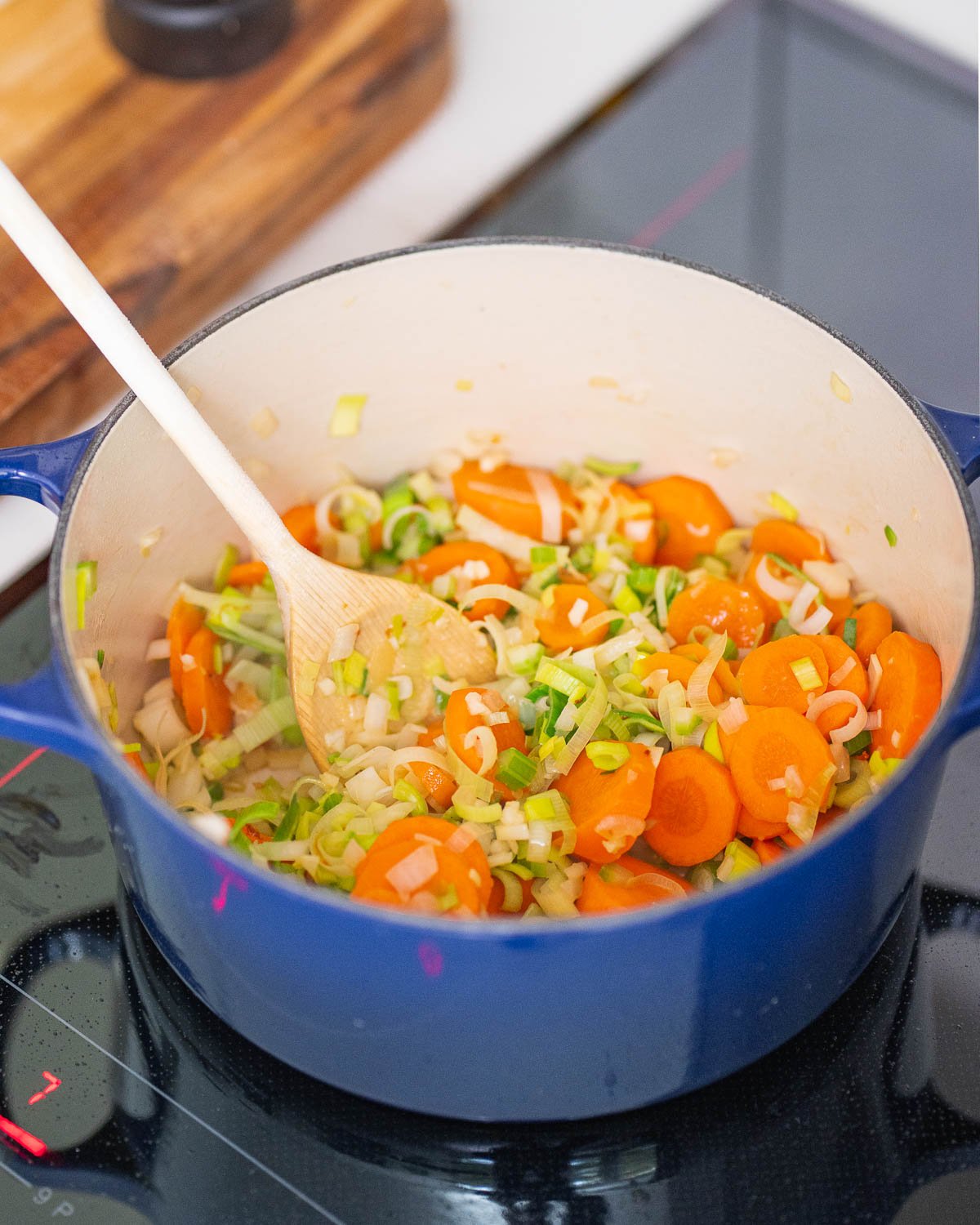 A blue saucepan on the stove holds sliced carrots, leeks and onions, which are stirred with a wooden spoon.