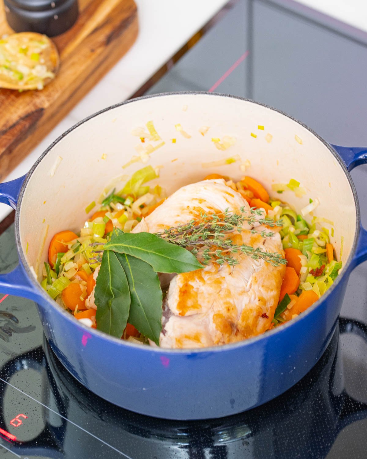 A blue pan on a stovetop containing a chicken breast, chopped vegetables, bay leaves and sprigs of thyme.