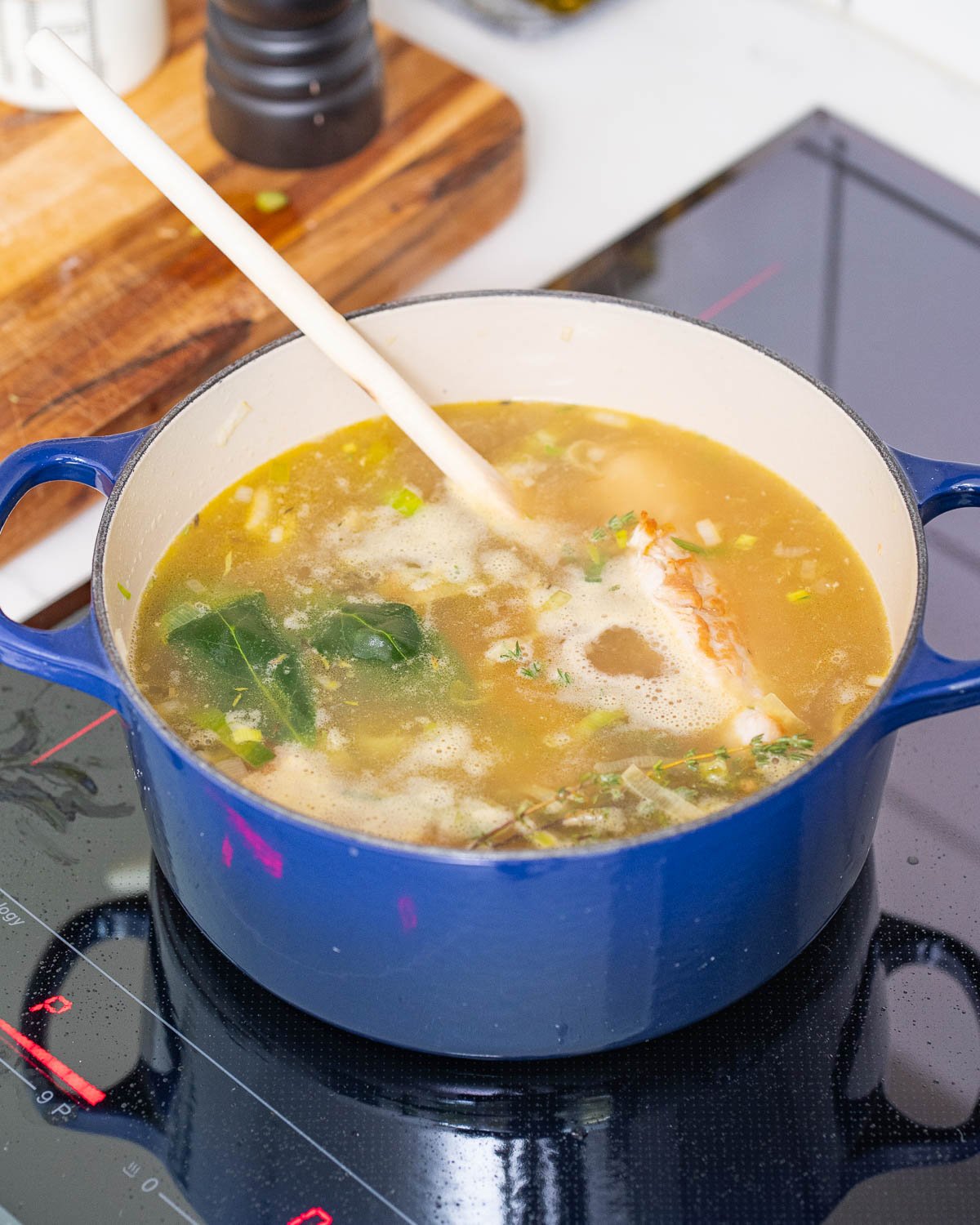 A blue pot filled with broth, vegetables and herbs, with a wooden spoon inside, sits on a stove.