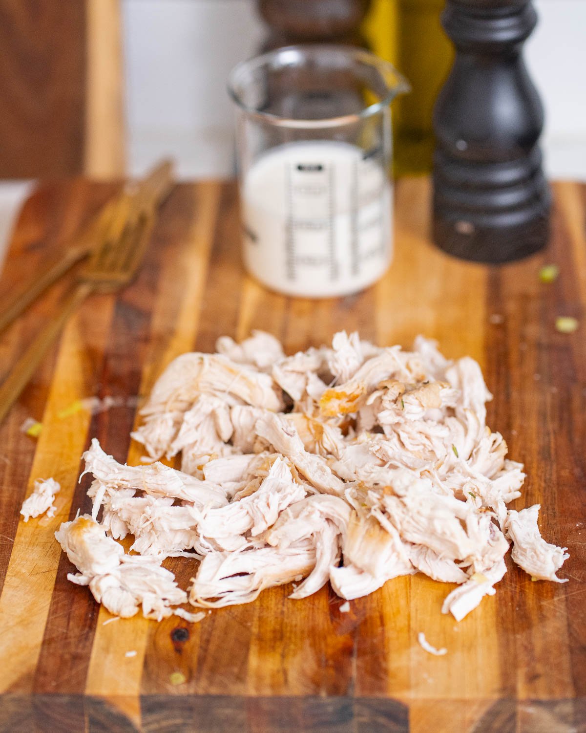 Shredded cooked chicken on a wooden cutting board, with a measuring glass of milk and a black pepper mill in the background.