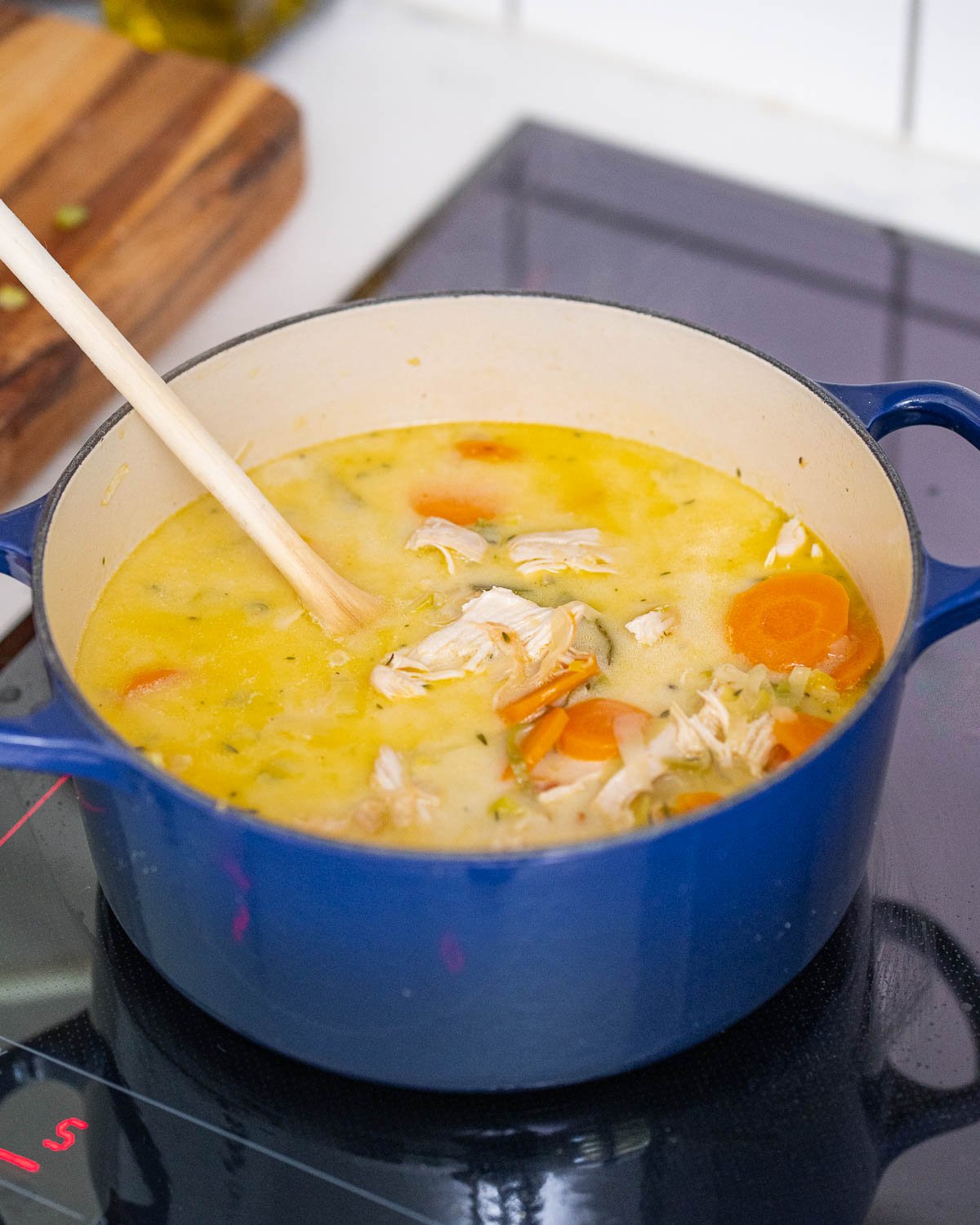 A blue saucepan on a stove containing chicken soup with carrots and a wooden spoon.