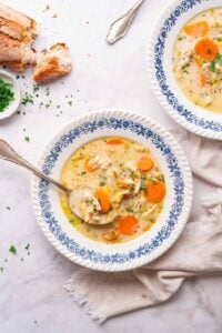 A bowl of chicken and carrot soup garnished with herbs, placed on a white table with a spoon and bread beside it.