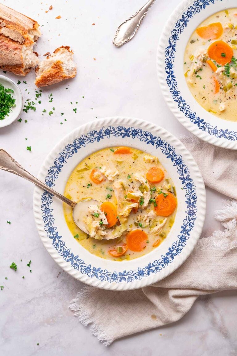 Un bol de soupe de poulet crémeuse aux légumes, ornée de carottes et d'herbes, repose à côté d'une cuillère en argent étincelante. Un autre bol et du pain fraîchement cuit sont visibles à proximité.