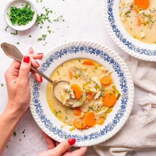 A person with red fingernails holds a spoon over a bowl of creamy vegetable chicken soup, rich in carrots and herbs. Nearby, on the immaculate white surface, another bowl sits next to a small dish of herbs and an additional spoon.