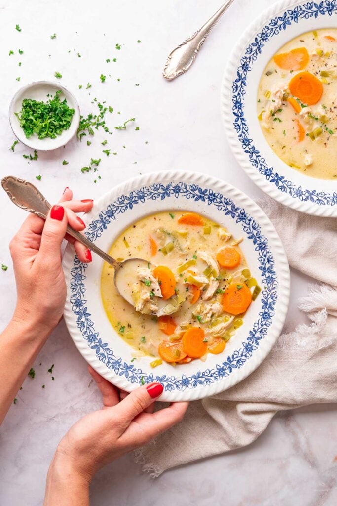 A person with red fingernails holds a spoon over a bowl of creamy vegetable chicken soup, rich in carrots and herbs. Nearby, on the immaculate white surface, another bowl sits next to a small dish of herbs and an additional spoon. 